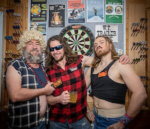 Three men in costume standing together in front of a dartboard. One wears a blonde, curly wig and a necklace of red plastic beads, another wears sunglasses and a checked shirt with the sleeves ripped off, and the third wears glittery eye shadow and has his vest tied up to expose his midriff
