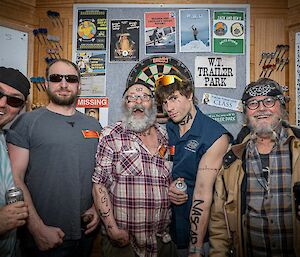 Five men in costume standing in front of a dartboard. A few are carrying beer cans, two are wearing sunglasses, and another two have drawn tattoo designs on their arms and faces with a permanent marker