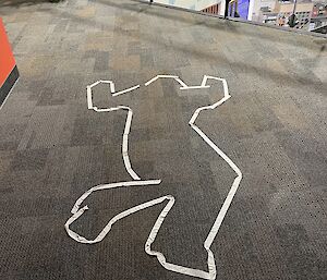 An imitation of a chalk outline of a dead body at a crime scene has been on a carpeted floor with white masking tape