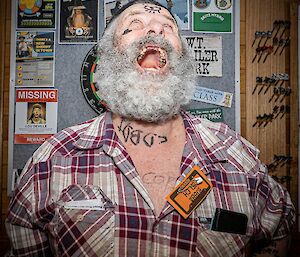 A man with a curly grey beard appears to be yelling at the ceiling. He wears a checked shirt with the top buttons undone, on which a name tag reading "Bubba Belch" is safety-pinned upside down. An eye has been drawn on his forehead with a marker