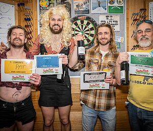 Four men in costume, standing in a row, smiling and holding up certificates and bottles of wine. One has tied up the waist of his shirt to expose his midriff, one wears a blonde wig and a woman's cocktail dress, one wears a flannel shirt and fake glasses, and one is holding up a wad of fake money