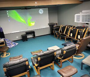 A lounge room with a large projector screen taking up most of the far wall. Armchairs and footstools are arranged in rows in front of the screen, with spare chairs stacked against one wall. There is a pile of quilts and cushions in one corner