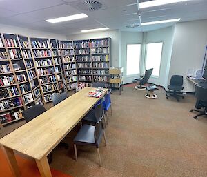 A room with one wall lined with tall shelves full of books, a long wooden study table down the centre, and some computer workstations against the wall opposite the bookshelves