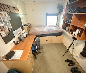 A single bedroom. The bed is at the far end, against a small, square window, with a system of storage drawers underneath. There are some storage shelves mounted on one wall, and a desk against the other. A pin-up board above the desk has some family photos and maps pinned to it