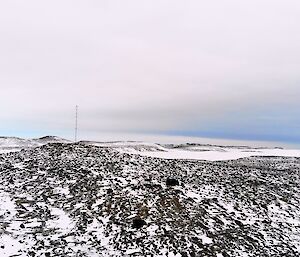 A sweeping aerial view of the brown rocks and snow of the Antarctic landscape.  A person can just me made out on the ground in the distance.