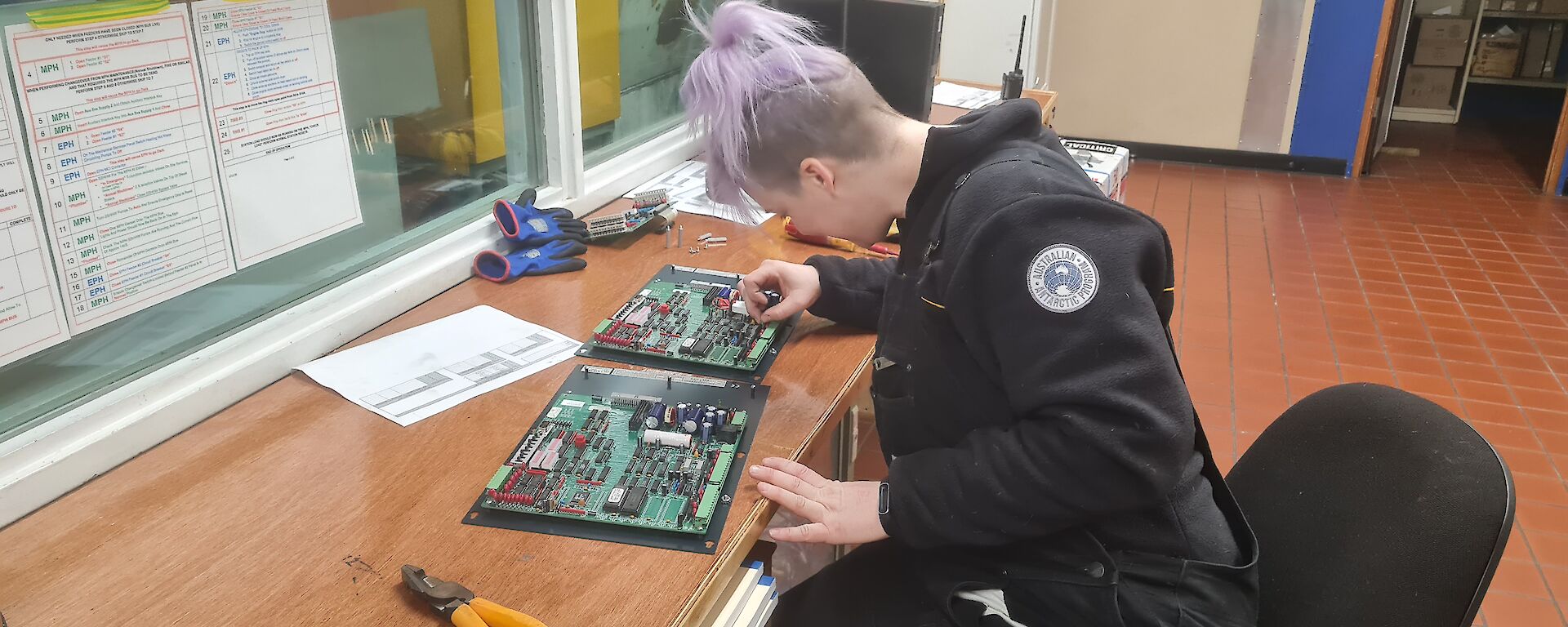 Woman with dyed purple hair sits at desk soldering electronics circuit boards