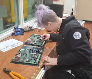 Woman with dyed purple hair sits at desk soldering electronics circuit boards