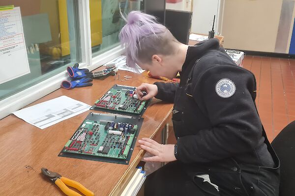 Woman with dyed purple hair sits at desk soldering electronics circuit boards