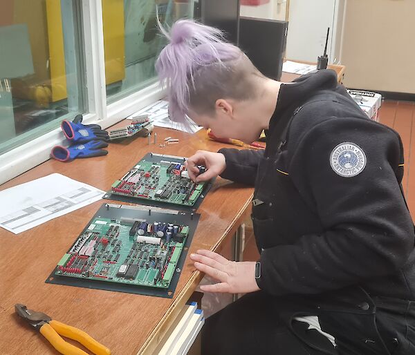 Woman with dyed purple hair sits at desk soldering electronics circuit boards