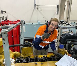 Woman stands over large diesel engine wearing PPE and giving thumbs up