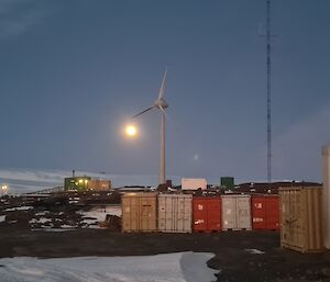 In foreground shipping containers, behind large wind turbine, and in back ground snow covered hills lit by a full moon.