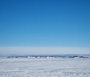 A landscape featuring flat, snowy ground textured with shallow sastrugi. In the distance is a line of low, black hills with grey and white layers of cloud just above them in an otherwise bright blue sky
