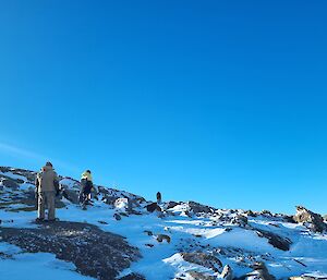 A view from the bottom of a snowy, rock-strewn slope. Three people are walking to the slope's summit carrying small pieces of equipment