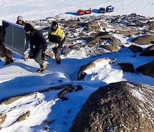 A view from a snowy, rock-strewn slope, looking down on four men, each holding the corner of a heavy metal frame with solar panels attached perpendicularly on one side, as they carry it up the slope
