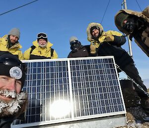 A selfie-angled photo of six people standing around the solar panels they have just installed in place by a radio mast on top of a hill. A man in the front, who is taking the photo, has a remarkable build-up of frost and small icicles in his beard and moustache