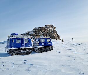 Rocks forming a small peak on top of a snow-covered slope. A dark blue Hägglunds vehicle is parked in front of the peak. Three people are exploring near the base of the peak