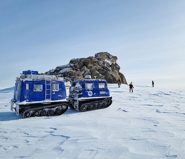 Rocks forming a small peak on top of a snow-covered slope. A dark blue Hägglunds vehicle is parked in front of the peak. Three people are exploring near the base of the peak