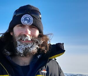 A picture of a man in a winter jacket and beanie, smiling at the camera. Condensation from his mouth and nose have formed frost and small icicles in his beard and moustache, including one especially prominent icicle hanging under his nose
