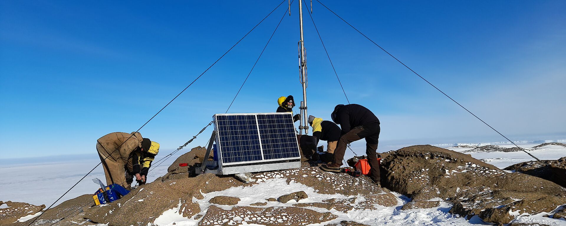 The top of a small rocky hill, dusted with snow, where a radio mast has been erected and anchored in place with four guy wires. Five men are working on securing the guy wires. A pair of solar panels mounted onto a metal frame sit nearby
