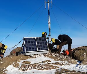The top of a small rocky hill, dusted with snow, where a radio mast has been erected and anchored in place with four guy wires. Five men are working on securing the guy wires. A pair of solar panels mounted onto a metal frame sit nearby