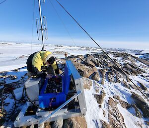 Beside a radio mast on top of a snow-covered, rocky hill, a man is bending over a blue chest fixed to a metal frame on the ground. The man is connecting the leads of an array of batteries inside the chest