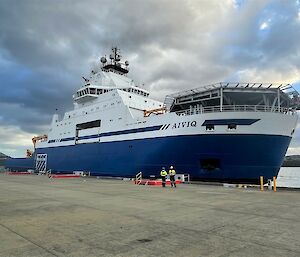 A blue and white ship with a helideck on one end moored at a large concrete dock.