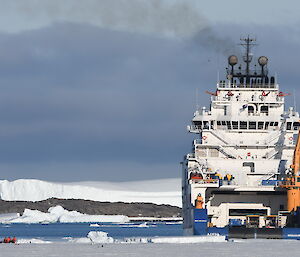 A blue and white ship sits in the sea ice. A group of expeditioners stand on the ice next to it.