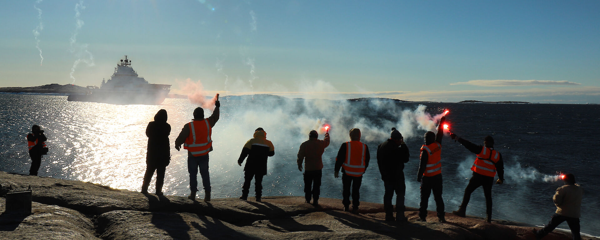 Ten people standing in a line on a rocky outcrop, with backs to camera and holding lit flares. They wave farewell to a ship in the distance.