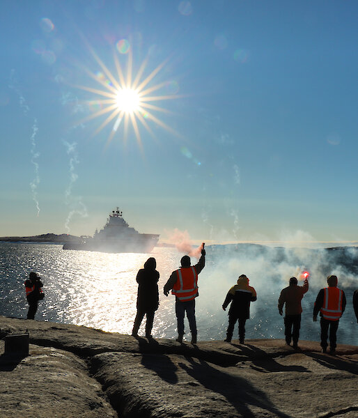 Ten people standing in a line on a rocky outcrop, with backs to camera and holding lit flares. They wave farewell to a ship in the distance.
