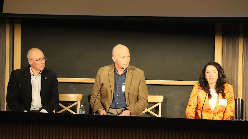 Two men and a woman seated at a discussion panel on stage. The woman is speaking into the microphone