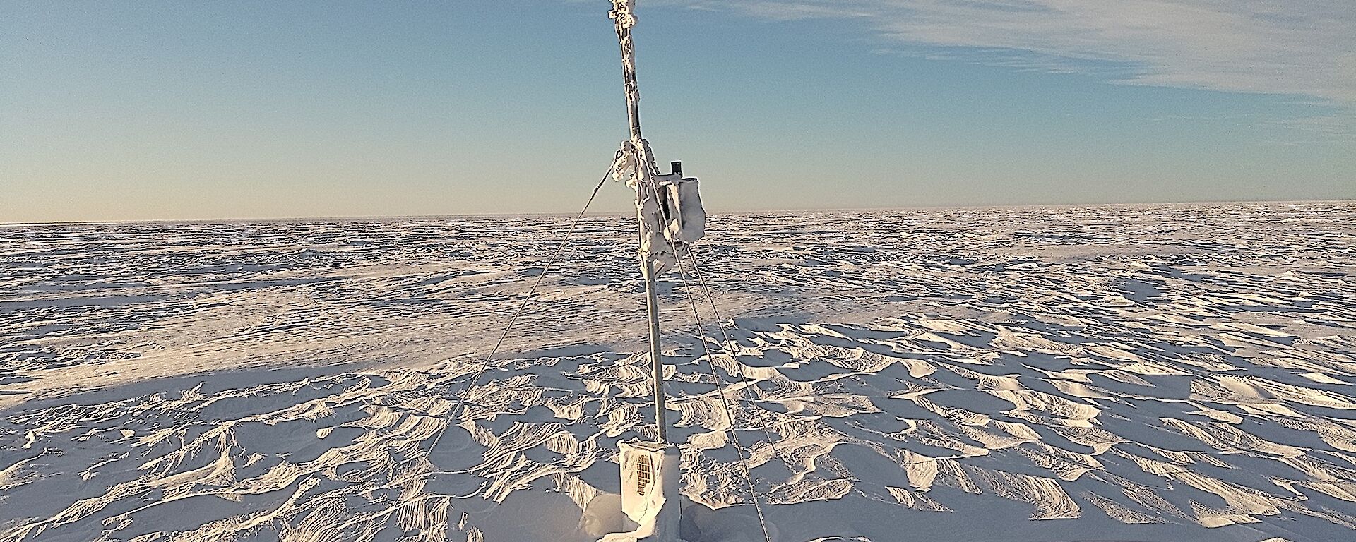 A wide plain covered with wind-rippled snow under a sky half-veiled with high cloud. A narrow mast with a wind vane and small propeller on top stands in the centre of the picture, apparently anchored to the ground with guy wires, but the base of the mast appears to be buried deep under the snow