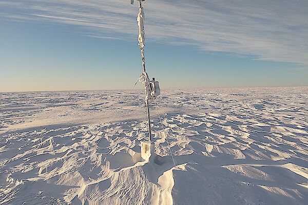 A wide plain covered with wind-rippled snow under a sky half-veiled with high cloud. A narrow mast with a wind vane and small propeller on top stands in the centre of the picture, apparently anchored to the ground with guy wires, but the base of the mast appears to be buried deep under the snow