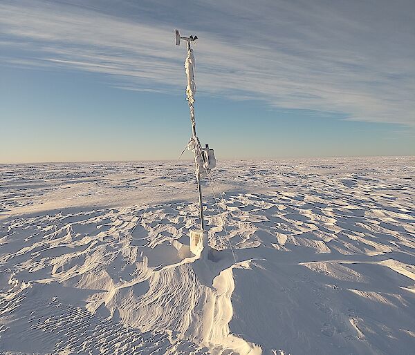 A wide plain covered with wind-rippled snow under a sky half-veiled with high cloud. A narrow mast with a wind vane and small propeller on top stands in the centre of the picture, apparently anchored to the ground with guy wires, but the base of the mast appears to be buried deep under the snow
