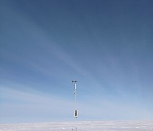 A narrow mast, bearing a solar panel halfway up and a wind vane with a small propeller on top, standing in a flat, snowy plain. Snow has been cleared to provide anchor points for guy wires attached to the mast