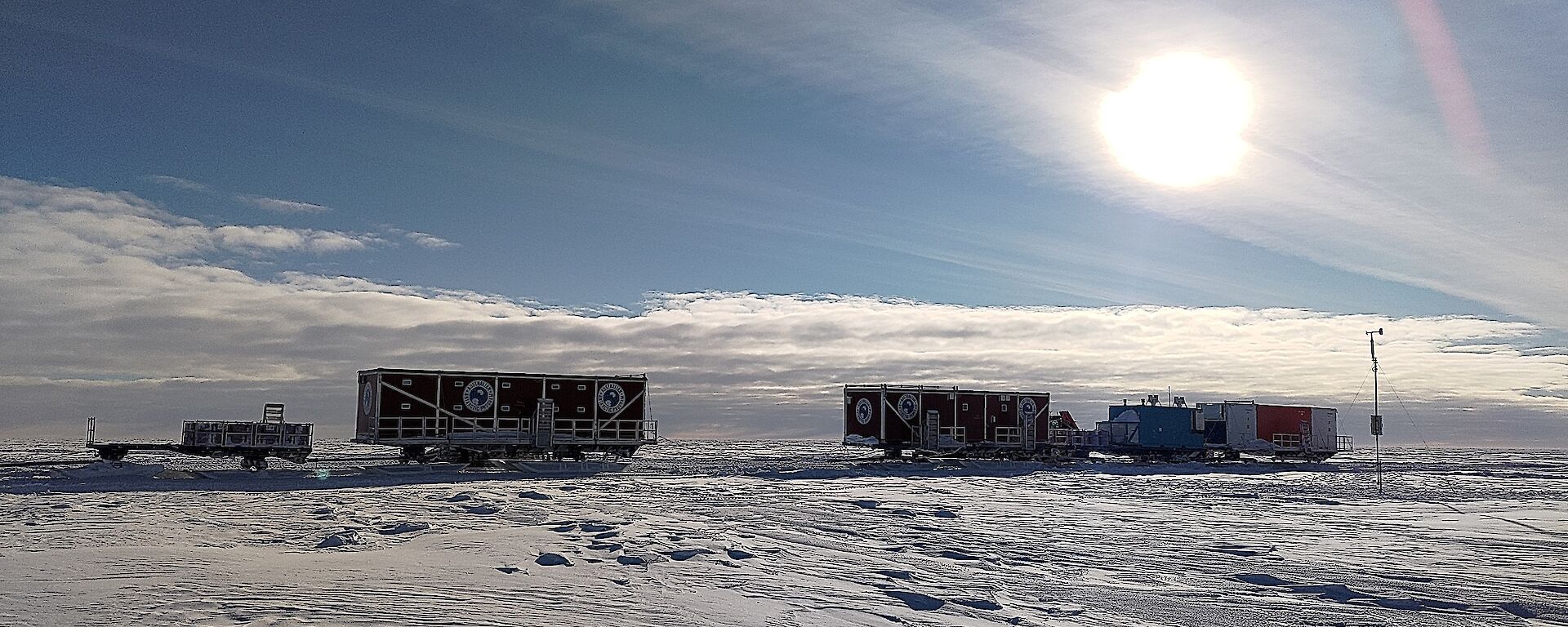 A series of cabins on sleds with a trailer at the rear, standing on a snowy plain near a narrow mast with a wind vane and a small propeller on top. The sun is shining brightly on the scene, slightly blurred behind a thin veil of high cloud
