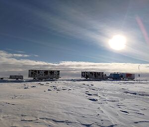A series of cabins on sleds with a trailer at the rear, standing on a snowy plain near a narrow mast with a wind vane and a small propeller on top. The sun is shining brightly on the scene, slightly blurred behind a thin veil of high cloud
