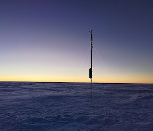 A snowy plain under a clear sky, with dawn's first light on the horizon. In the centre of the picture is the newly-erected Automatic Weather Station: a narrow cylindrical unit with a wind vane and small propeller on top, mounted on top of a narrow mast with a small solar panel attached to it, all fixed to the ground with three guy wires