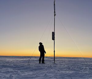 A snowy plain under a clear sky, with dawn's first light on the horizon. In the centre of the picture is the newly-erected Automatic Weather Station. A man stands beside the mast looking up at the unit on the top, appearing in silhouette against the golden glow of dawn