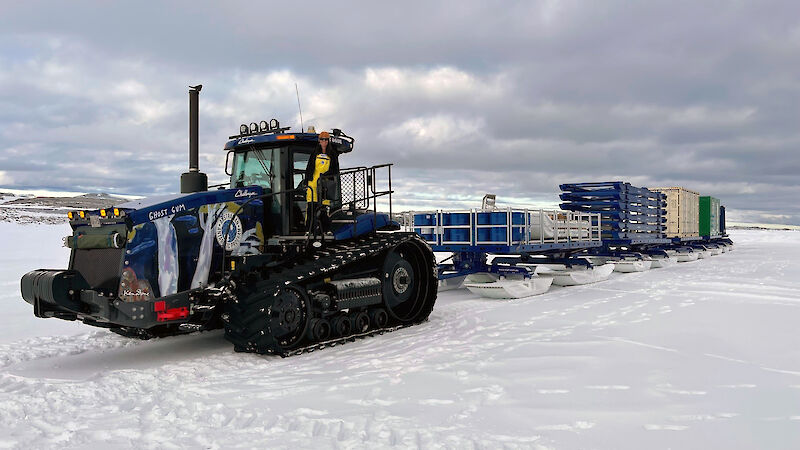 A blue tractor with sleds behind it on the ice in Antarctica. A woman stands in the door of the tractor.