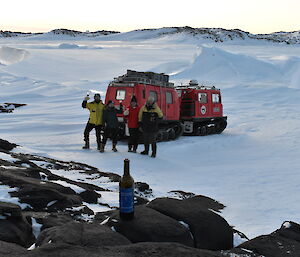 Four people in front of red Hägglunds with a bottle of wine sitting on the rocks in front of them