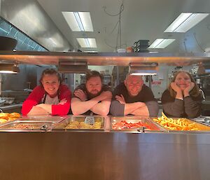 Four smiling people behind a bain marie of food