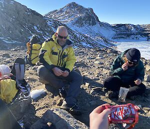 Two people sitting on the rocky ground eating chocolate bars for energy in the cold