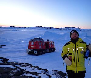 A man in a yellow jacket holding a bottle of wine in front of a red Hägglunds in the snow