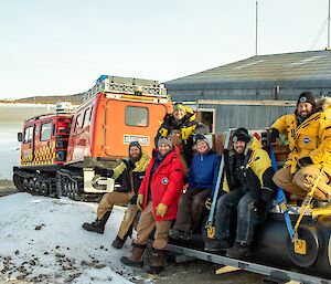 Group of six expeditioners sit of sled being towed behind red Hägglunds vehicle