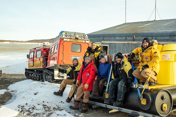 Group of six expeditioners sit of sled being towed behind red Hägglunds vehicle