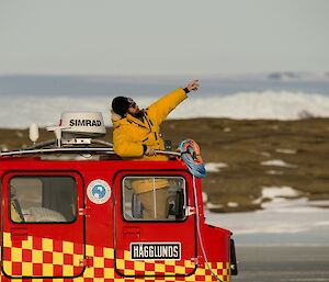 Man standing with head and shoulders out of the top 'sunroof' of the Hägglunds points off into the distance