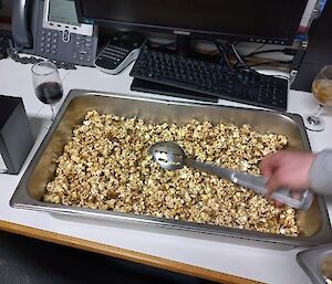 A large bain-marie container of popcorn. An arm can been seen scooping out some popcorn with a large serving spoon.