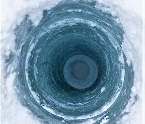 A close-up view into a hole drilled into lake ice. The top of the hole is bordered with powdery white snow, while the icy interior is a deep aquamarine colour