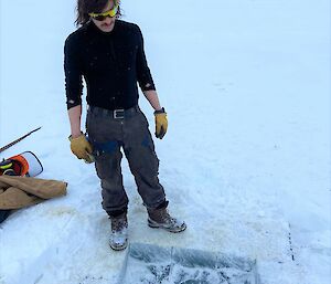 A man in snow-laden work boots and reflective sunglasses is looking down at a hole that has been drilled into the thick ice layer on top of a lake. The frozen lake surface he stands on is covered thickly with snow