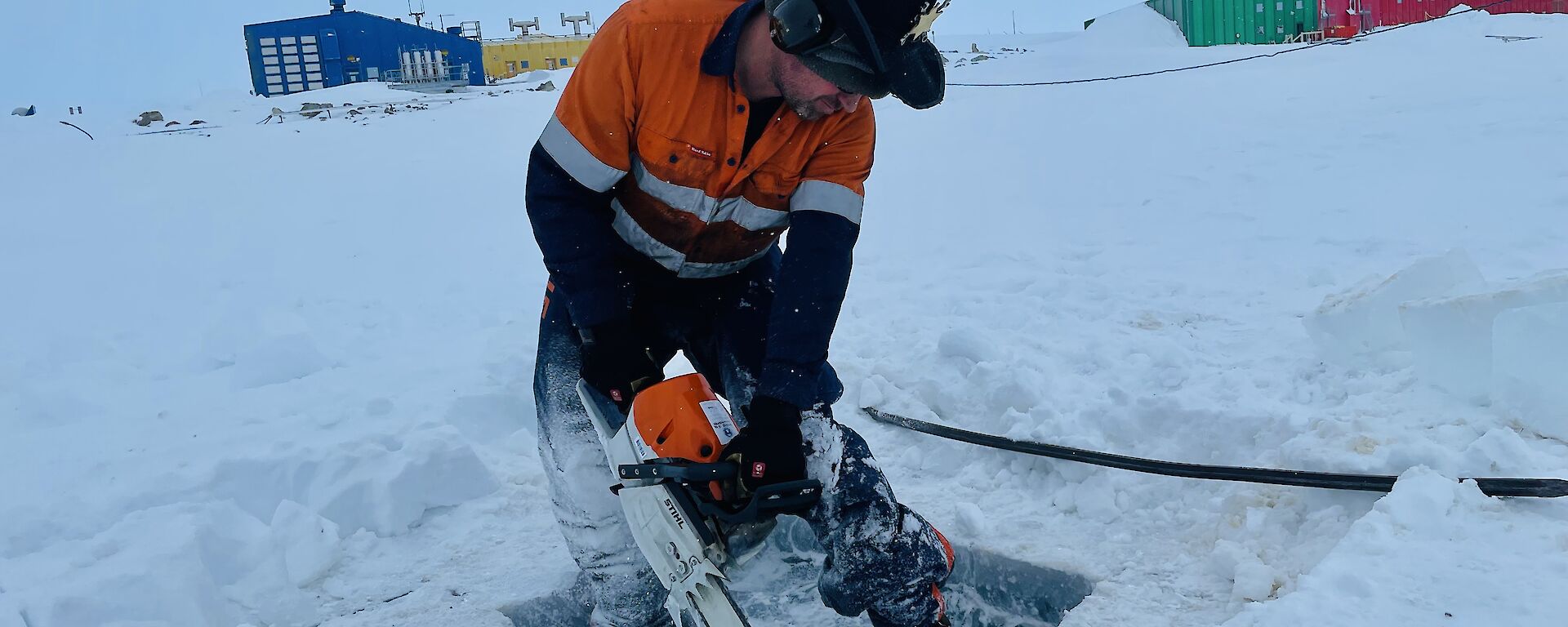 A man wearing high-vis work clothing, earmuffs and a cowboy hat is operating a chainsaw to cut through a thick sheet of ice covering a lake. Behind him, across a stretch of snow-covered ground, are the bright blue, yellow, green and red buildings of an Antarctic station